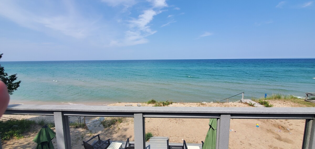 The image captures a tranquil view of Lake Michigan's expansive blue waters, bordered by a sandy beach. The horizon meets the sky, showcasing gentle waves. A wooden deck and green seating are visible in the foreground, inviting relaxation by the water.
