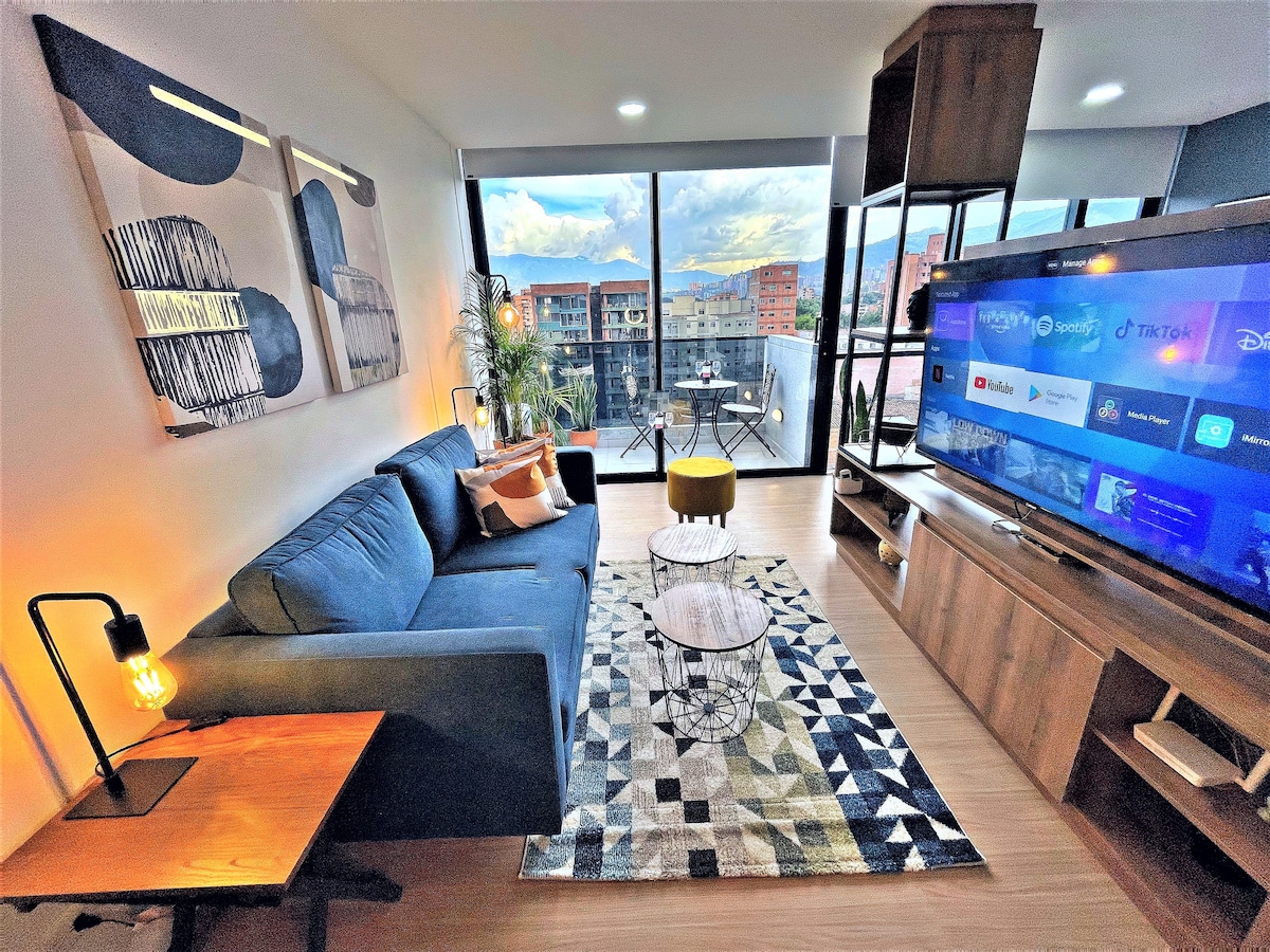 A modern living area features a comfortable blue sofa and a patterned area rug. A sleek television stands adjacent to a large window that offers expansive city views, complemented by a small balcony with a bistro table and chairs. Decorative art pieces and a warm lamp add subtle touches.