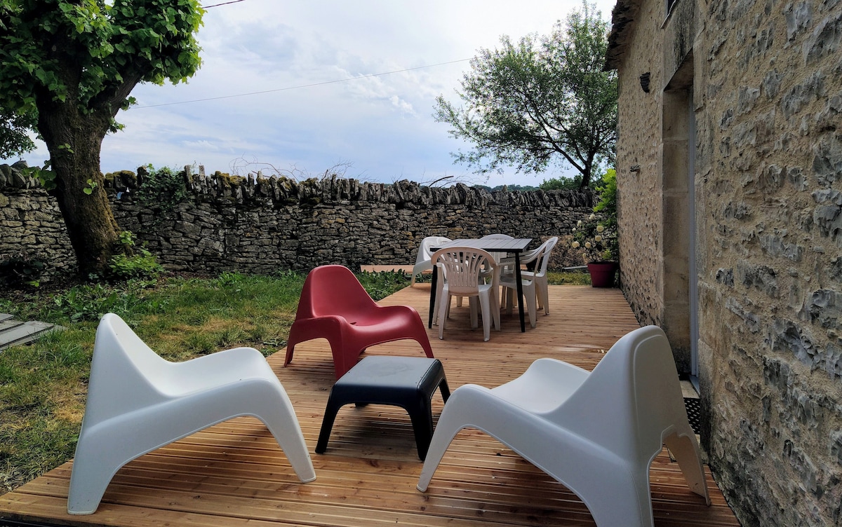 An outdoor deck area features a combination of plastic lounge chairs in red and white, alongside a black table. A rustic stone wall is visible in the background, and greenery from nearby trees enhances the natural setting.