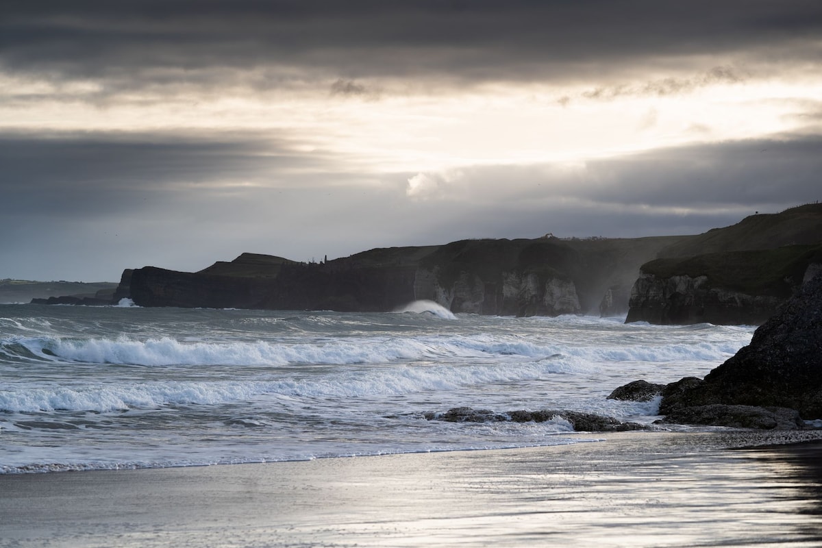 The coastline is displayed with rolling waves, meeting a sandy shore under a moody sky. Dark cliffs can be seen in the distance, partially shrouded in mist, while the water reflects the soft, muted light of the evening.