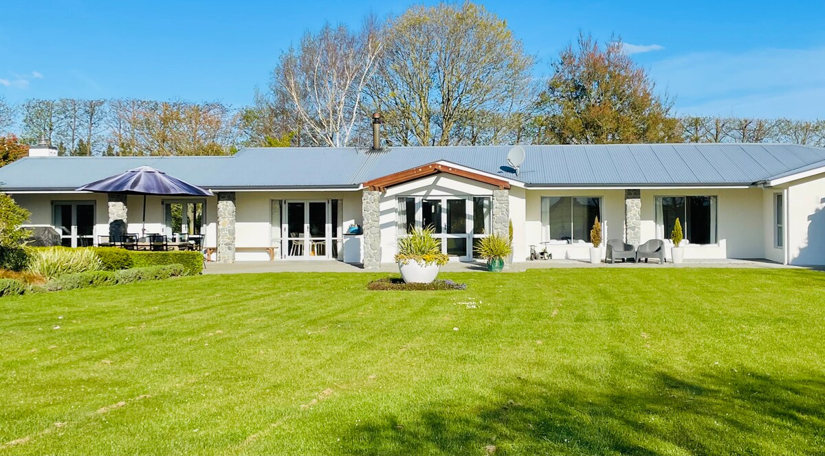 The exterior of the homestead is presented with a well-maintained lawn and a spacious patio, featuring outdoor seating and an umbrella. Large windows allow natural light to fill the interior, surrounded by greenery and trees, enhancing the serene country setting.