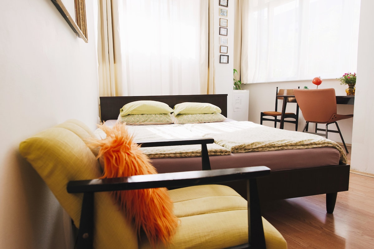 A bright bedroom is illuminated by natural light streaming through large windows. The spacious bed is adorned with soft linens, while a cozy chair with an orange accent pillow provides a comfortable seating area. A small desk is visible in the corner, accompanied by a potted plant.