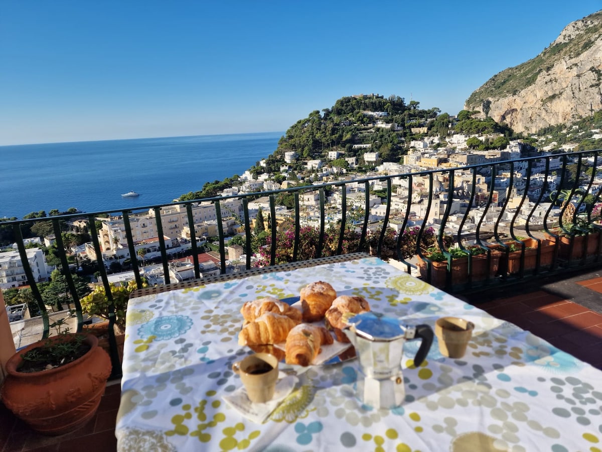 A table is set on a terrace overlooking the sea, featuring a coffee pot and baked goods. The scenic backdrop includes lush hills and vibrant buildings cascading down towards the coastline under a clear blue sky.