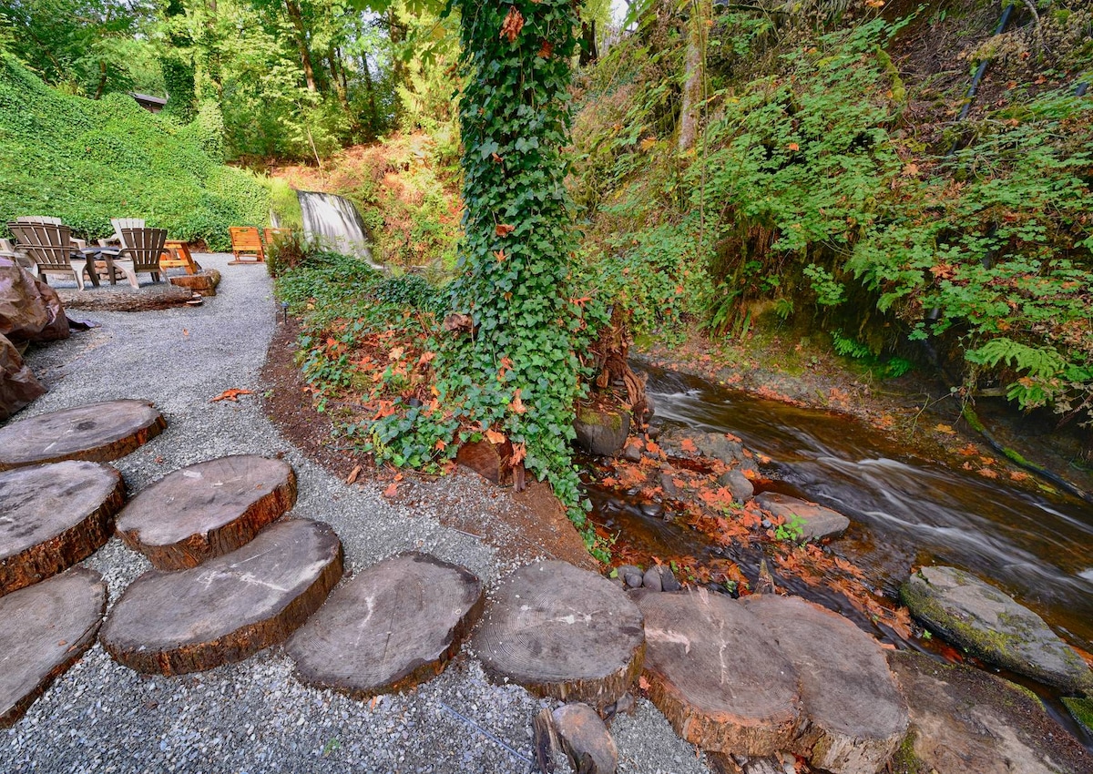 A serene outdoor area features a pebbled path lined with circular wooden stepping stones. A gentle creek flows beside it, accompanied by lush greenery and a small waterfall in the background. Rustic seating is arranged nearby for relaxation in nature.