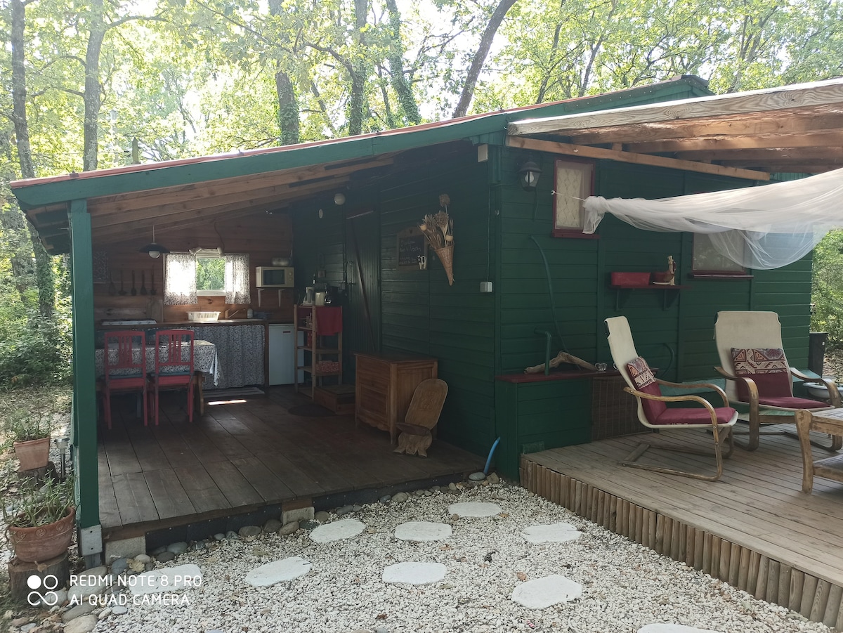 The chalet is presented in a natural setting, featuring a shaded porch with two chairs and a small side table. The exterior showcases wooden siding and a sloped roof. Within view, a kitchen area and dining space are observable through open doors.