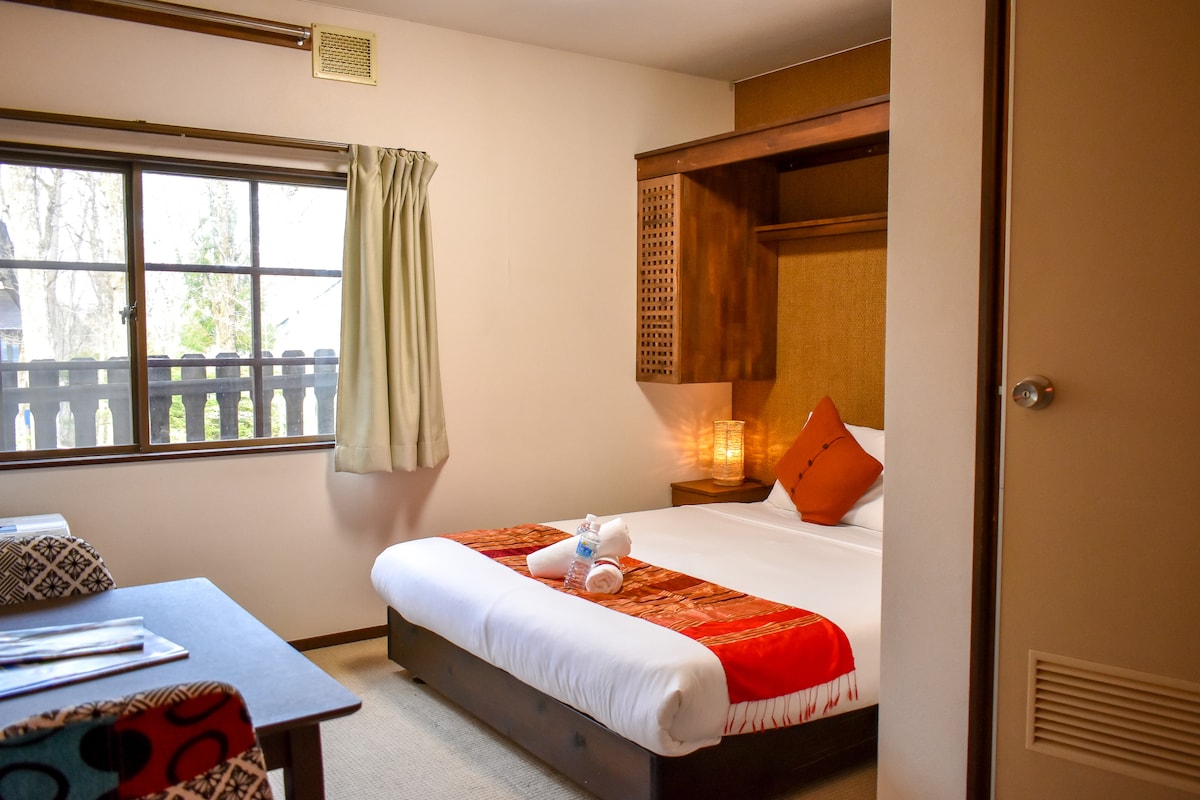 A compact bedroom features a spacious bed with a red and white comforter. A cozy sitting area with a wooden table is visible, along with a window framed by light curtains that allow natural light to brighten the room.