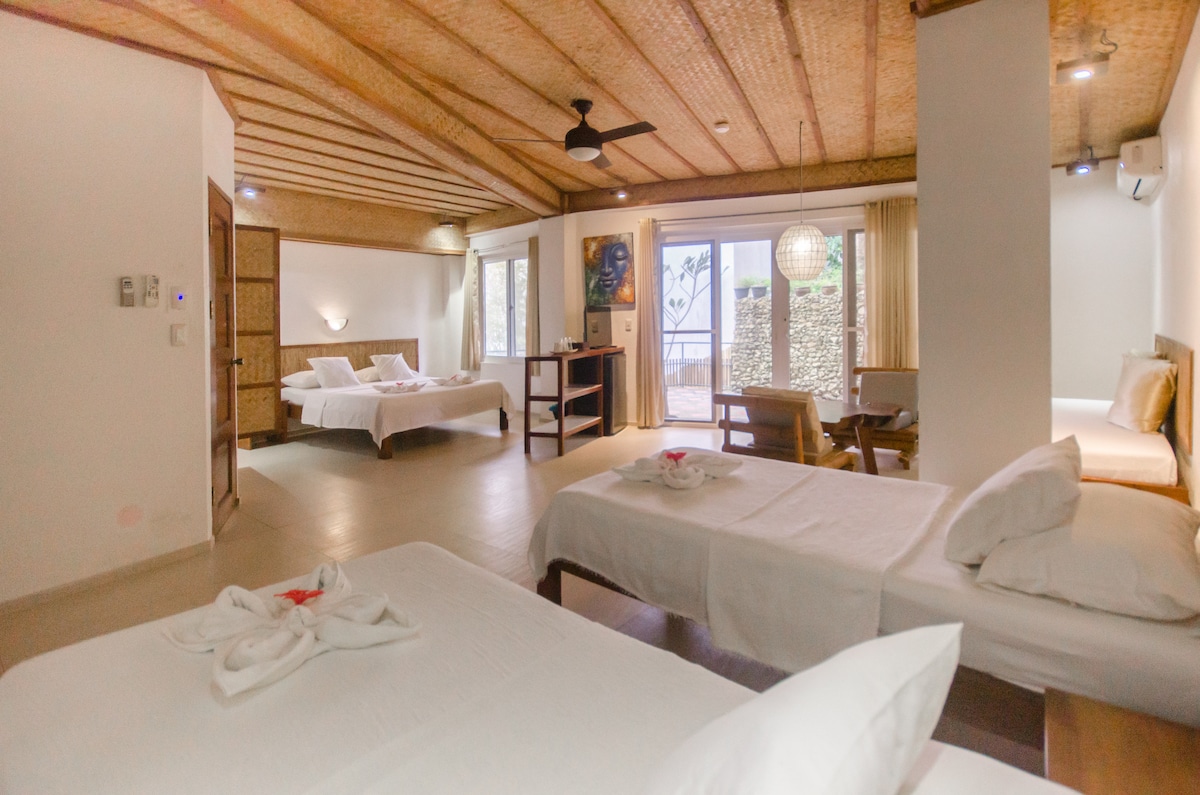 The spacious family room features two single beds and one queen-sized bed, adorned with fresh linens. Large windows allow natural light to fill the room, with a private balcony visible in the background. A comfortable seating area and decorative elements enhance the inviting ambiance.
