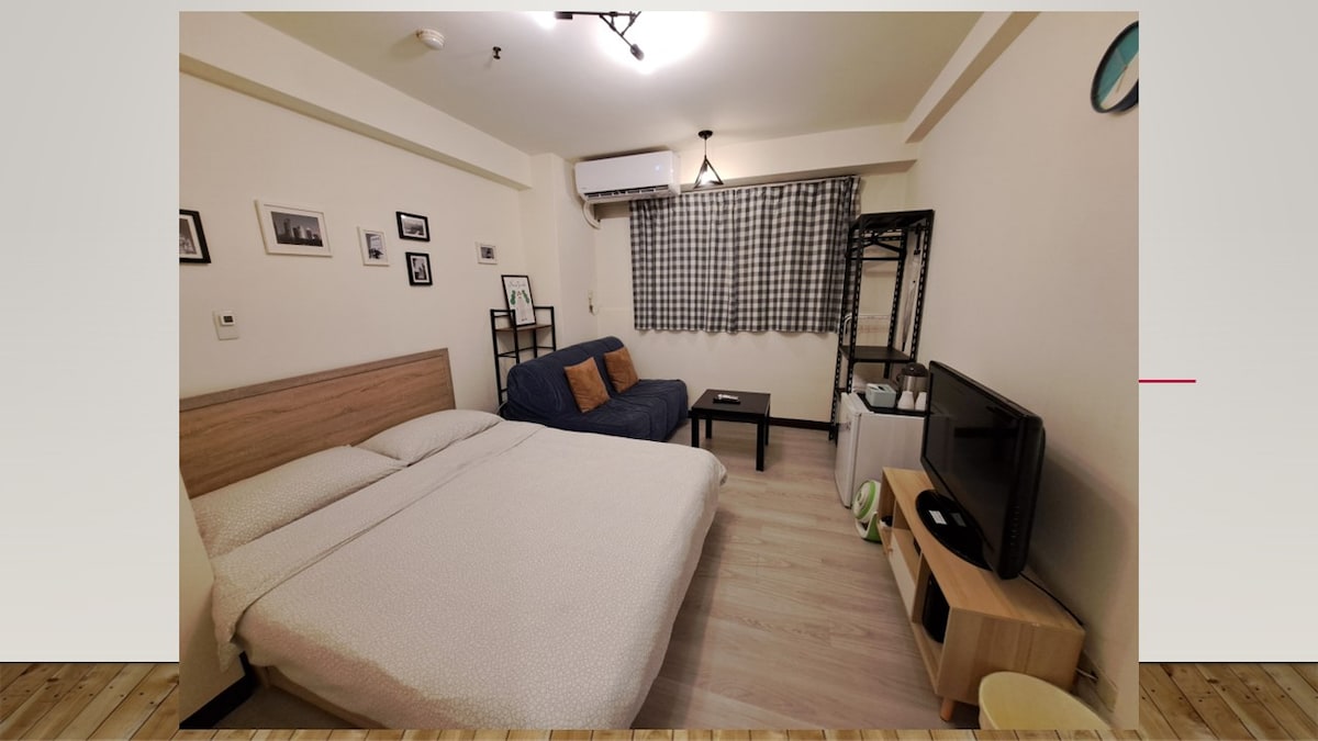 A cozy room features a spacious bed and a sofa for relaxation. A television is positioned on a light wooden stand, accompanied by a small coffee table. Natural light filters through a window adorned with checked curtains, while framed photographs add character to the wall.