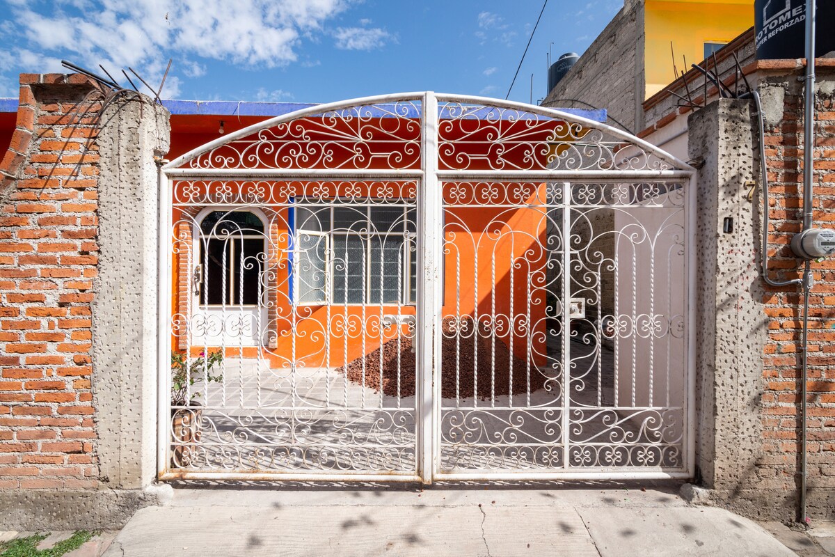 An ornate white wrought iron gate features intricate designs, leading to a welcoming entrance. The facade showcases vibrant orange walls, complemented by a large window and a pathway that invites guests into the property. Surrounding greenery adds a touch of nature to the space.
