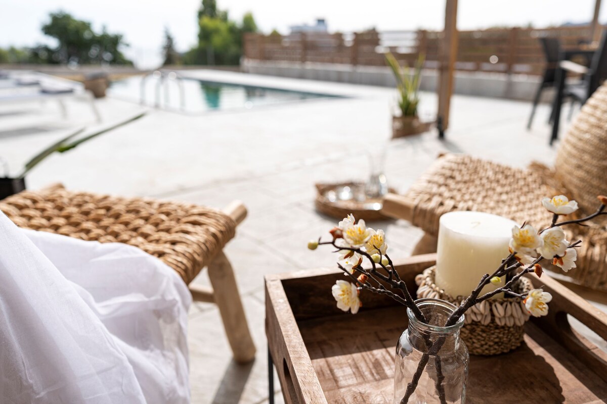 A wooden tray is presented with a candle and a small vase containing delicate flowers. The background includes a private infinity pool and sun loungers, creating a serene outdoor space.