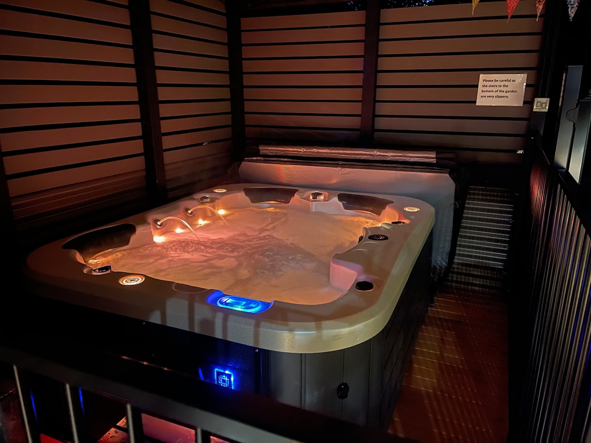 A luxurious hydrotherapy spa is presented, nestled within a fenced area. Soft lighting enhances the relaxing atmosphere, with controls illuminated around the edges. The spa offers a spacious design, inviting guests to unwind in comfort.
