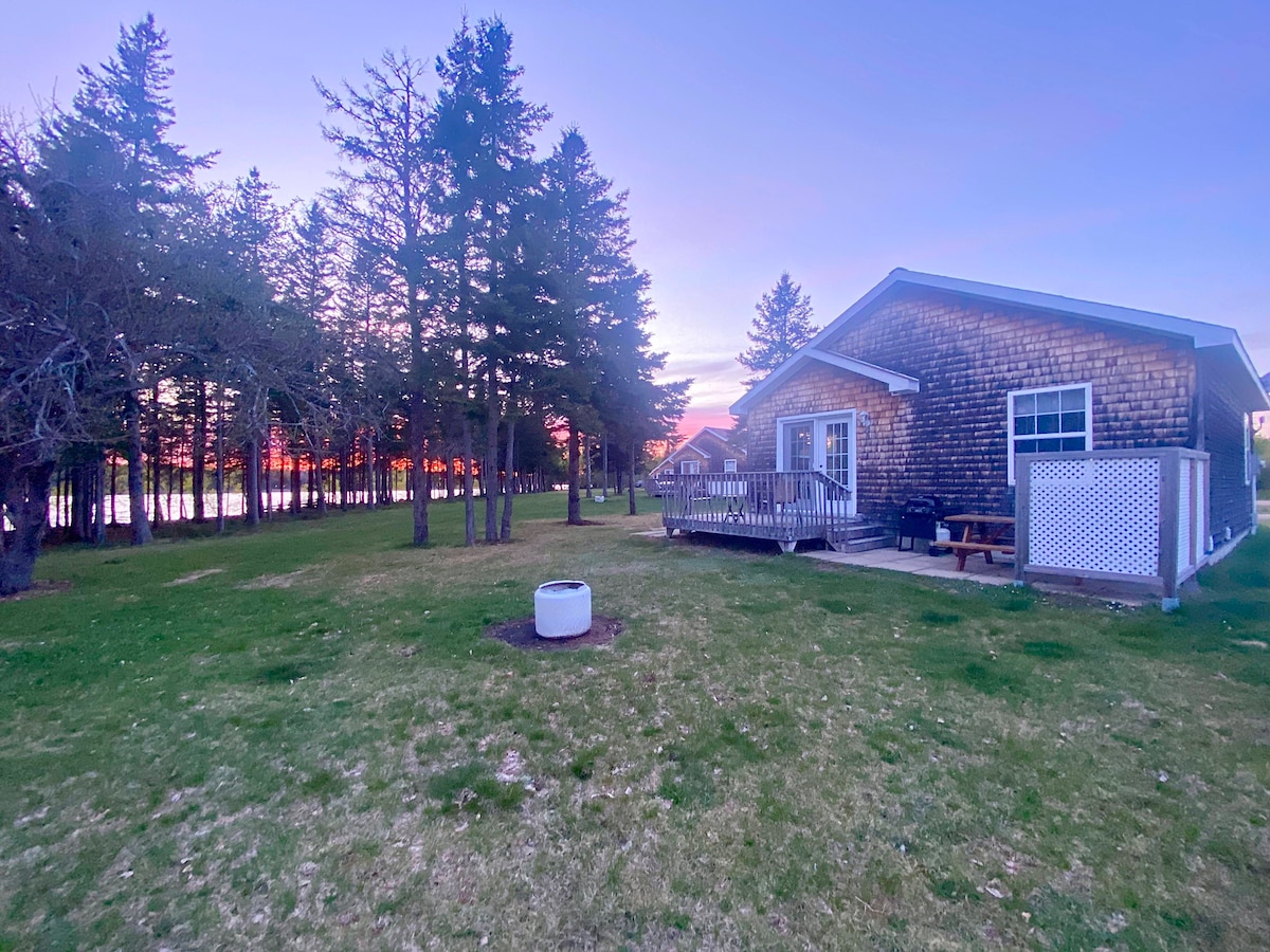 A cottage is set amidst a lush green yard, bordered by tall trees. The side deck features a seating area, while a fire pit with chairs is situated in the backyard. A colorful sunset casts a warm glow over the scene.