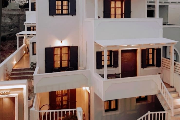 A two-story white building showcases a traditional design with dark blue shutters and warm lighting. A covered balcony provides seating for two, while the entrance features decorative details, creating a welcoming appearance against the evening backdrop.