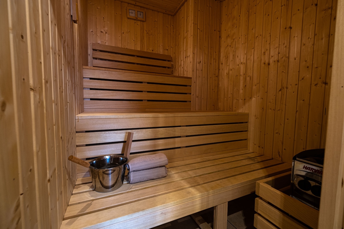 A wooden sauna interior features stacked benches against warm wood paneling. A metal bucket and wooden ladle are placed on the lower bench, while neatly rolled towels rest beside them. Subtle lighting enhances the serene atmosphere of this relaxing space.