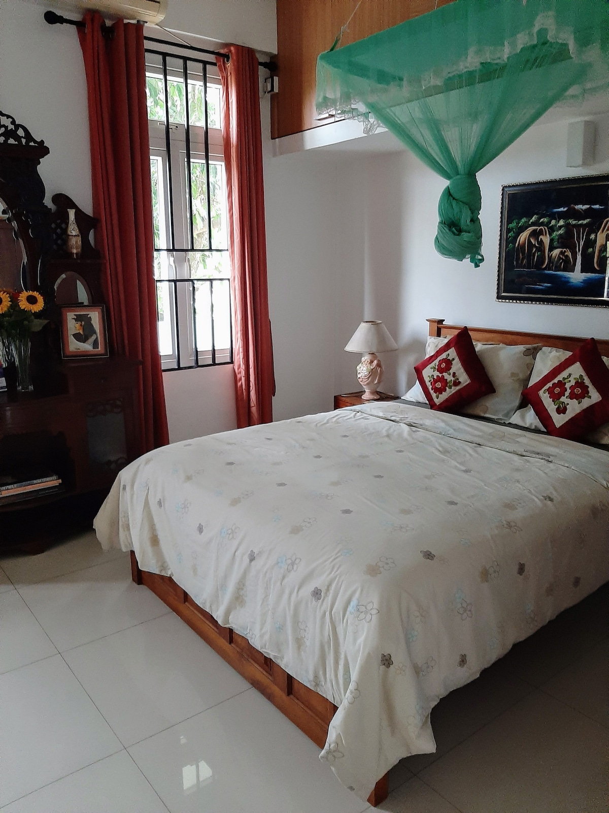 A cozy room features a queen bed adorned with decorative pillows and a light-colored quilt. Natural light enters through a window with protective bars, complemented by orange drapes. A bedside lamp and a small wooden display with flowers enhance the space's charm.