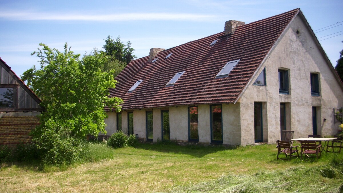 A spacious, rustic building features a sloping roof with multiple skylights. Large windows allow natural light to fill the interior, while a tree and outdoor seating create a welcoming space on the grassy grounds.