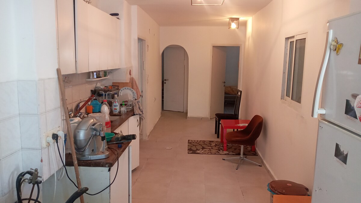 A spacious kitchen area features light-colored cabinets and a countertop with appliances arranged. A dining space contains a red table and a chair. An archway leads to other rooms, and a neutral tile floor extends throughout the area, maintaining a clean and functional look.