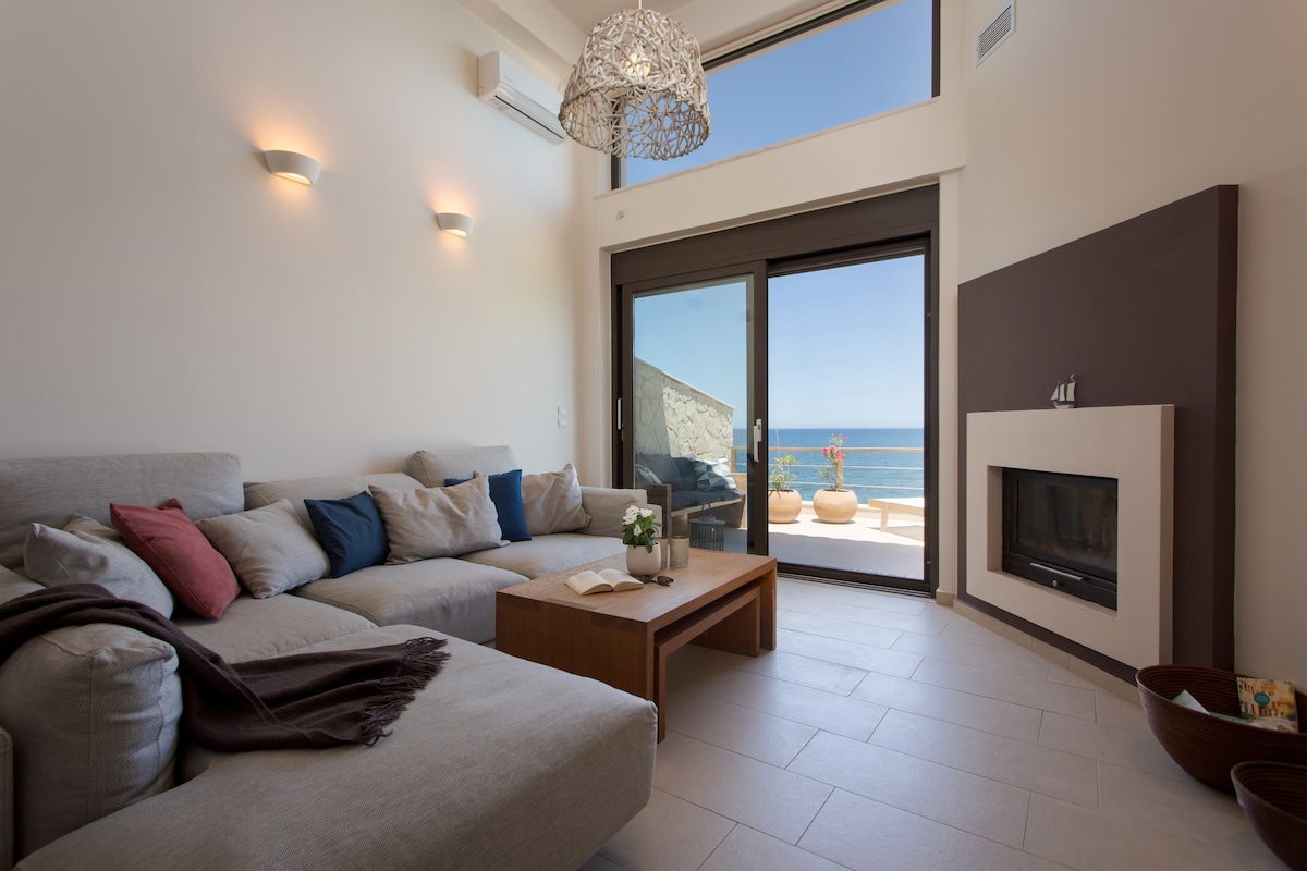 A spacious living area features a large corner sofa adorned with decorative pillows, complemented by a wooden coffee table. Natural light floods through glass doors that offer views of the sea. A cozy fireplace enhances the room's comfort, while light fixtures provide warm illumination.