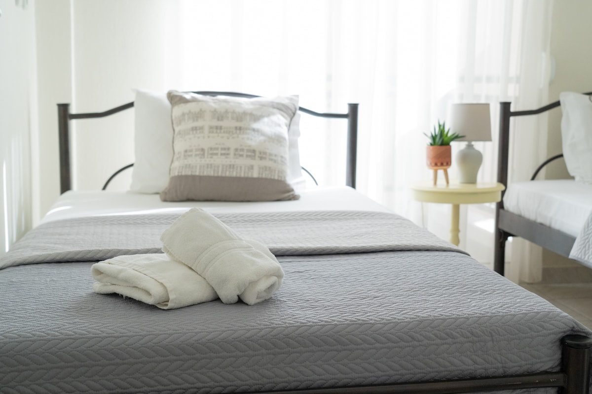 A bright bedroom features a spacious bed with crisp linens and a decorative pillow. Fresh towels are neatly folded on the bed. A side table holds a small lamp and a potted plant, with natural light streaming through sheer curtains.