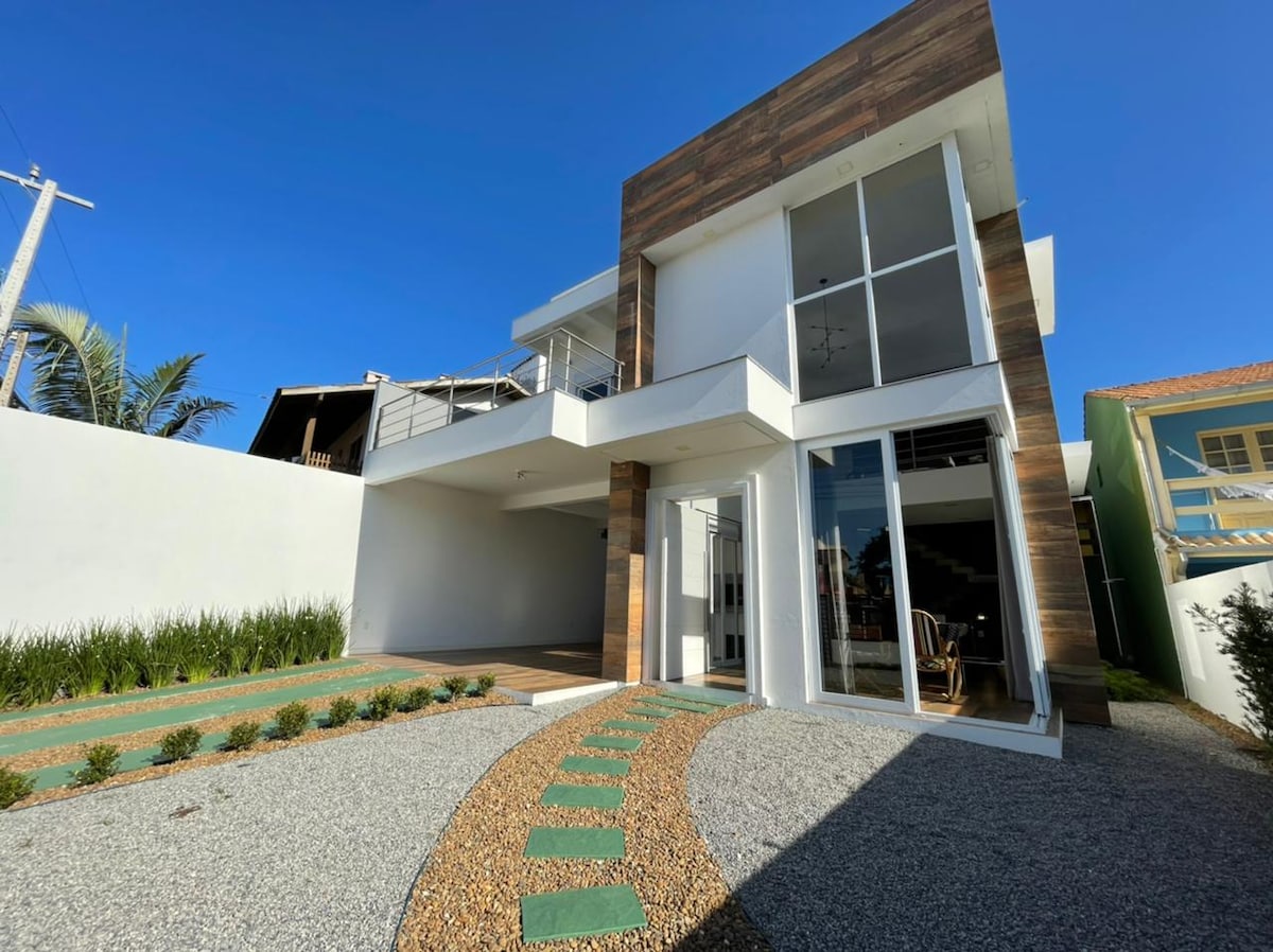 A modern two-story house features large glass windows and wooden accents. The entrance is approached via a stone pathway bordered by well-maintained greenery. An outdoor area is visible, enhancing the contemporary design of the home.