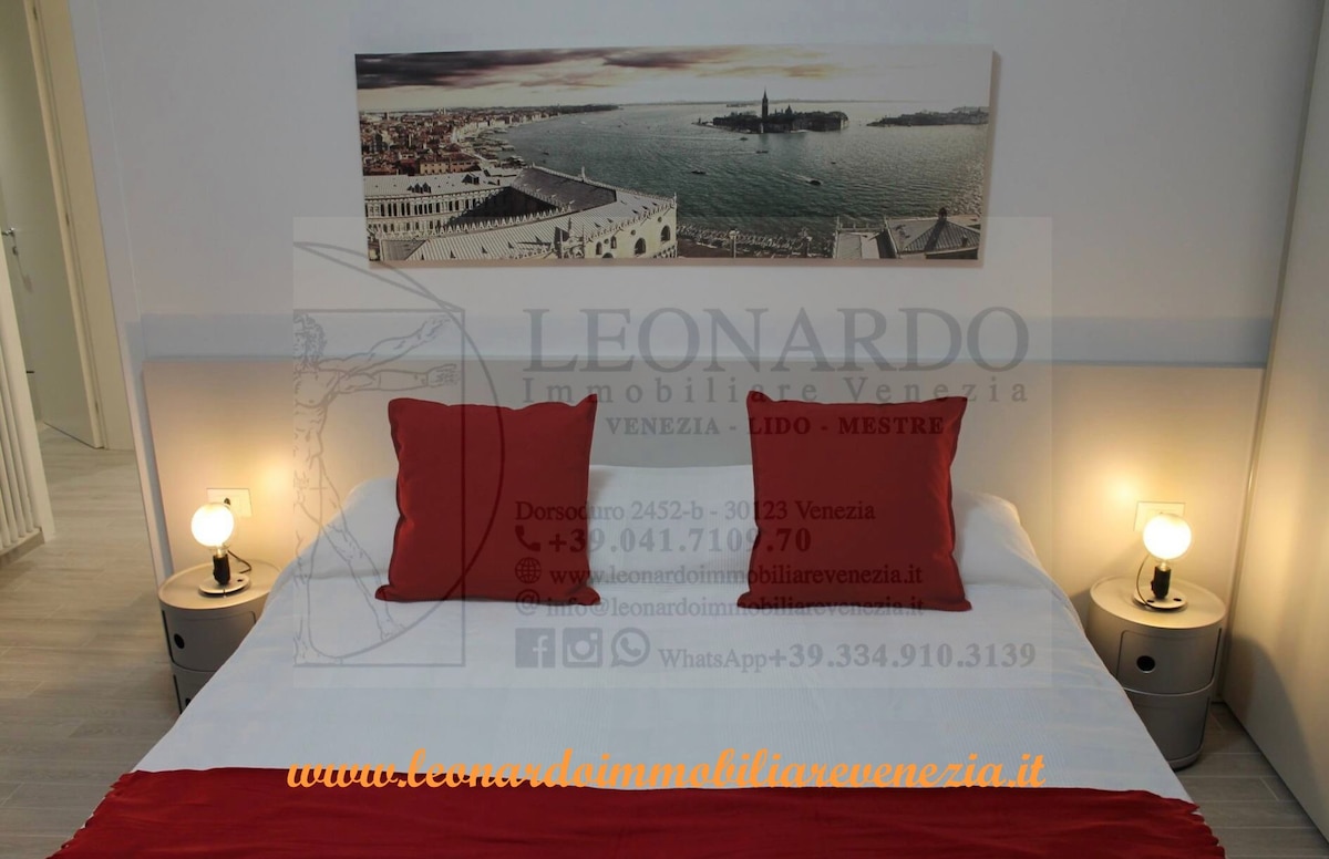 A spacious bed is draped with crisp white linens, flanked by two bedside lamps that provide soft illumination. Red accent pillows add a touch of color. A large scenic wall print showcases a view of Venice, complementing the calming atmosphere of the room.