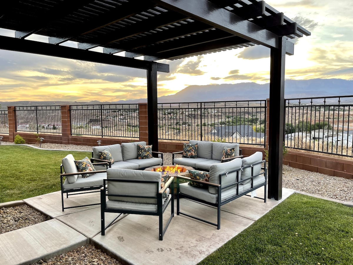 An outdoor seating area is arranged around a fire pit, under a shaded pergola. Comfortable couches with floral cushions are positioned on a stone patio, with scenic views of the sunset and distant mountains visible beyond the fence.