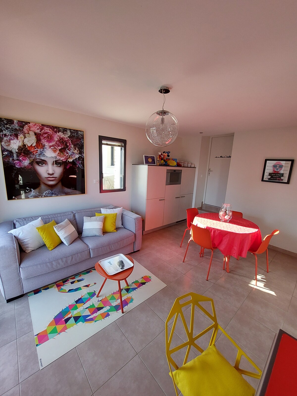 A bright living area features a light gray sofa adorned with colorful cushions, complemented by a round red dining table surrounded by orange chairs. A playful area rug adds interest to the tiled floor, while a large floral artwork decorates the wall, providing a touch of color.