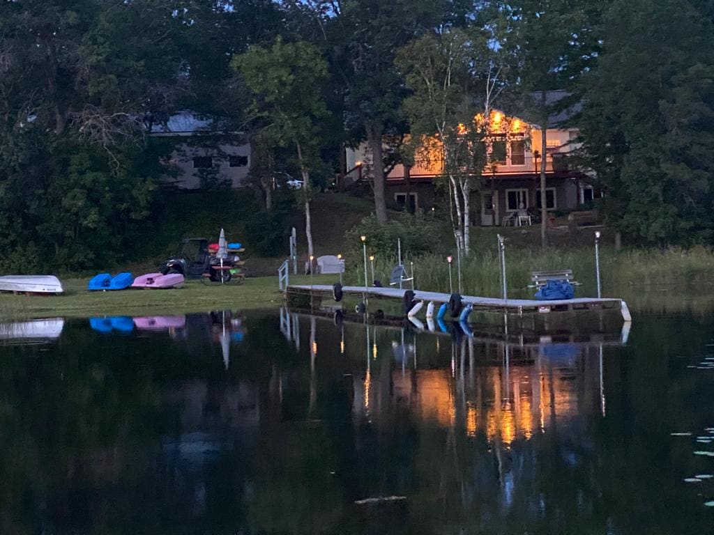 The image captures a tranquil lake scene at dusk, with the reflection of a well-lit home visible on the water's surface. A dock, equipped with kayaks and paddle boats, is situated at the lake's edge, surrounded by lush greenery.