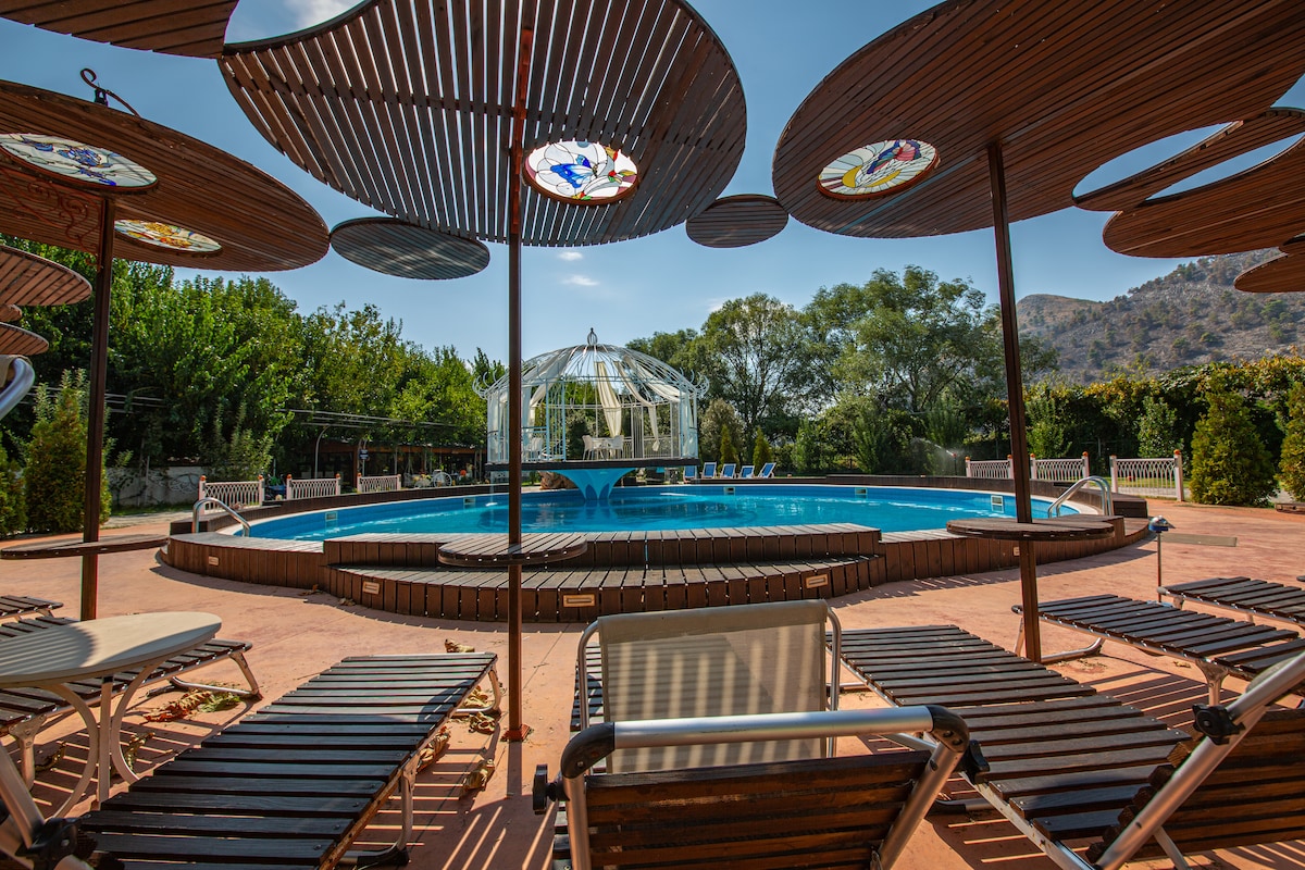 A spacious swimming pool is surrounded by a wooden deck, featuring several lounge chairs arranged for relaxation. Decorative umbrellas with stained glass accents provide shade over select seating areas, while lush greenery and trees frame the pool, creating a serene environment.