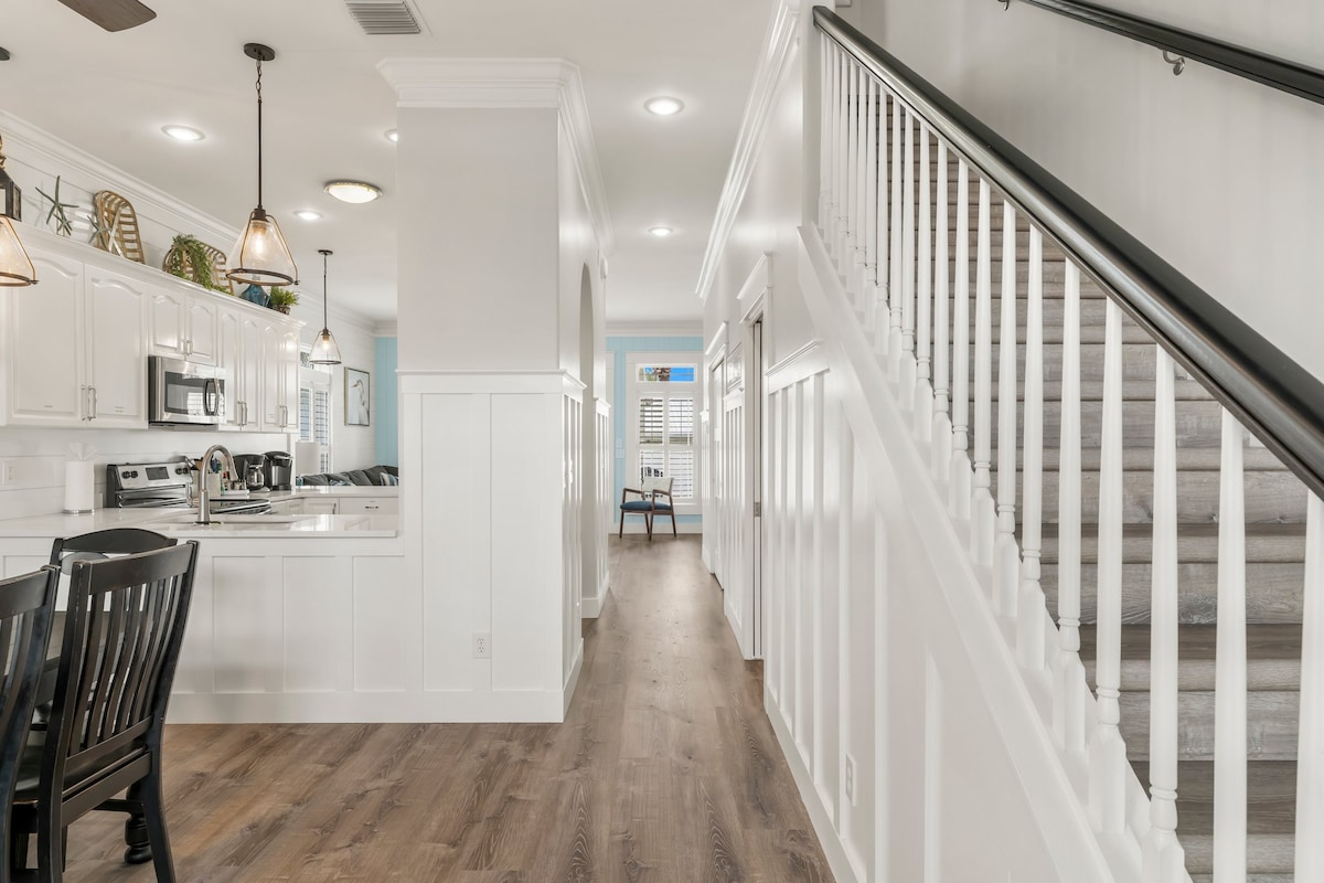 A bright and airy hallway features light-colored walls and wooden flooring. To the left, a fully equipped kitchen is visible, along with a dining area featuring dark wood chairs. A staircase leads to the upper level, adding to the spacious feel of the home.