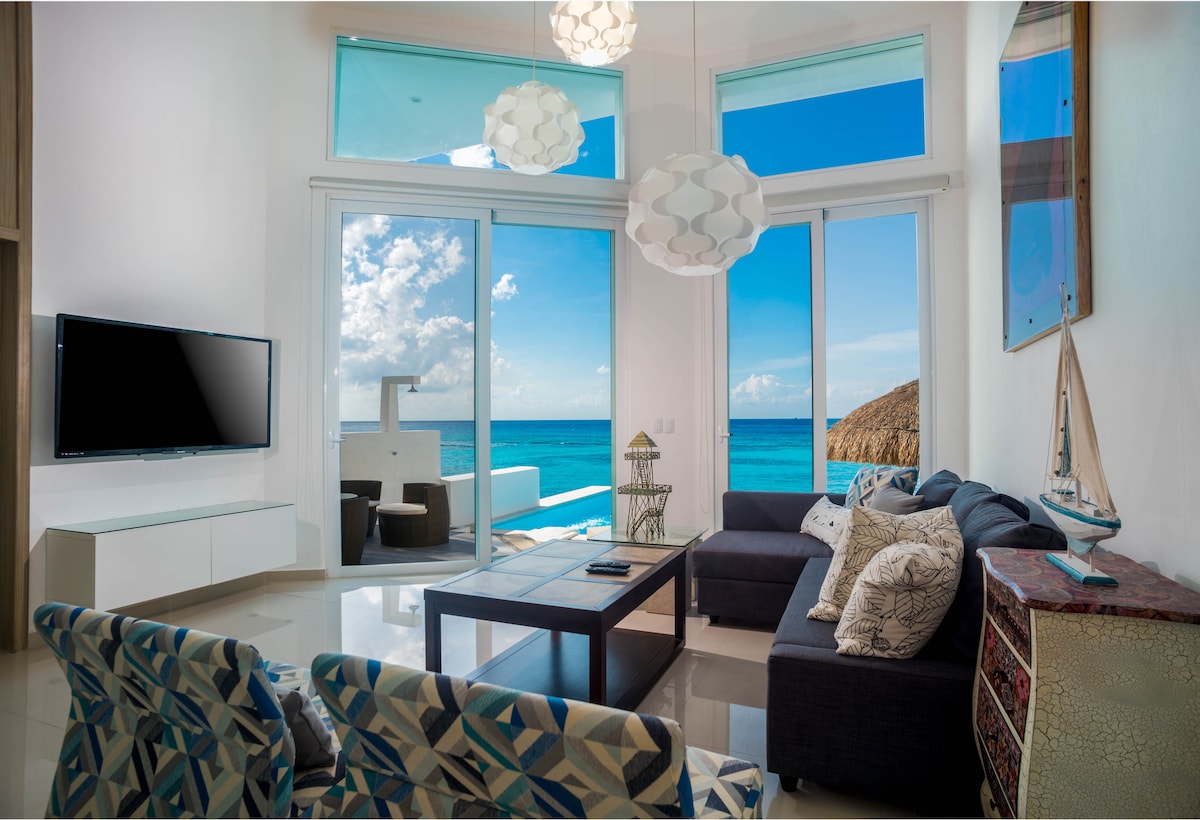 A bright and airy living area is highlighted by large sliding glass doors offering ocean views. Comfortable seating arrangements include patterned chairs and a sectional sofa. Natural light floods the space, complemented by modern pendant lighting. A flat-screen TV is mounted on the wall opposite the seating.