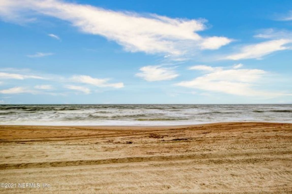 A serene beach scene is presented with soft, sandy shores stretching toward the gentle waves of the Atlantic Ocean. A clear blue sky dotted with wispy clouds creates a calm atmosphere, highlighting the natural beauty of Amelia Island.