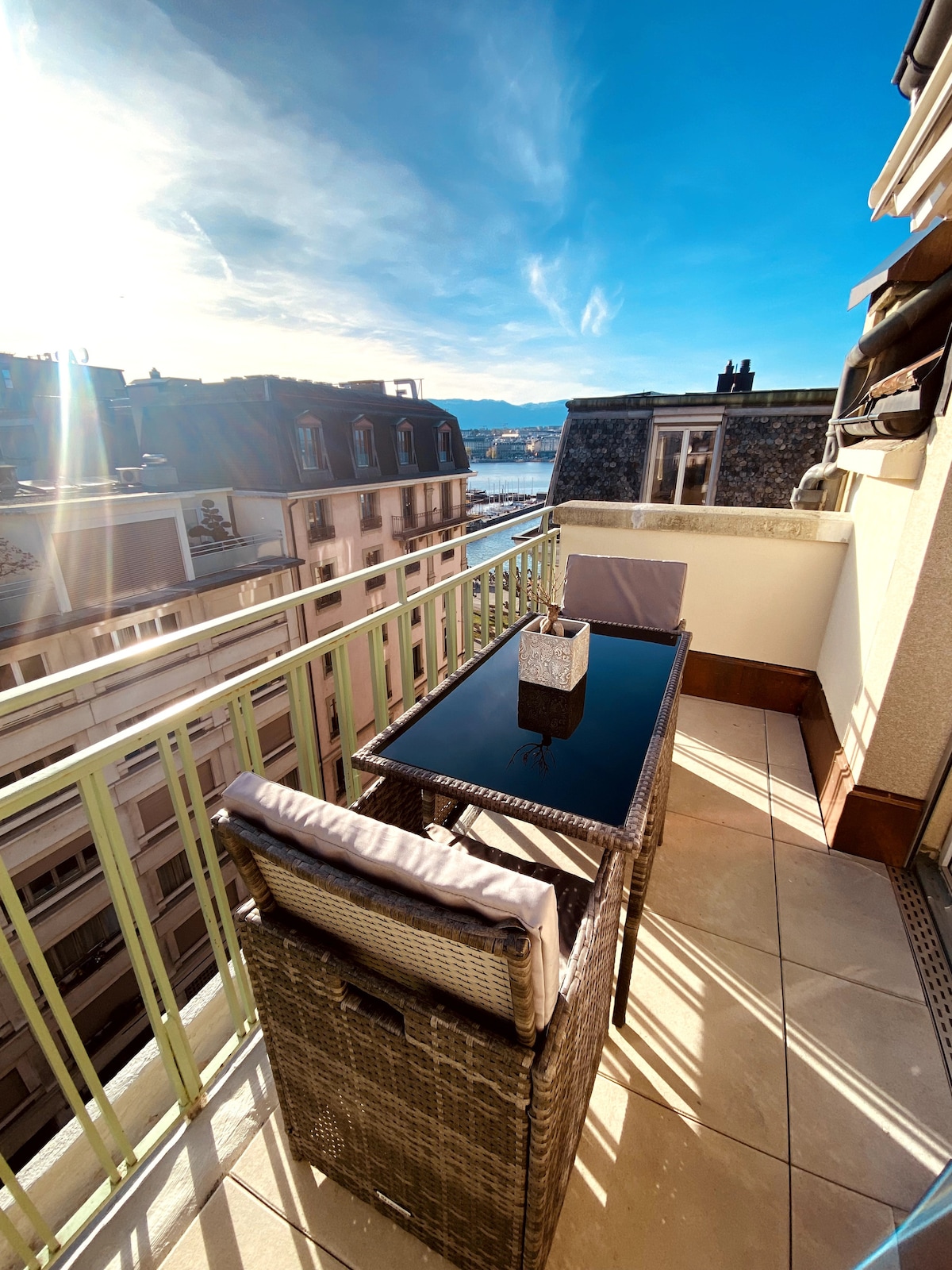 A balcony is displayed with a rectangular glass-top table and two wicker chairs. The area is surrounded by a light green railing, offering views of neighboring rooftops and blue skies. Sunlight reflects off the glass table, creating a bright atmosphere.