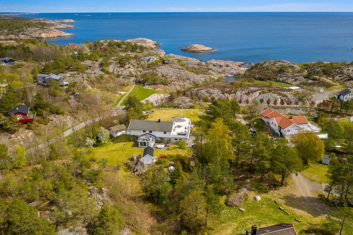 The aerial view showcases a spacious summer house surrounded by lush greenery and rocky landscapes. The ocean is visible in the background, with islands dotting the horizon. Nearby, other houses blend into the natural setting, highlighting the tranquil environment.