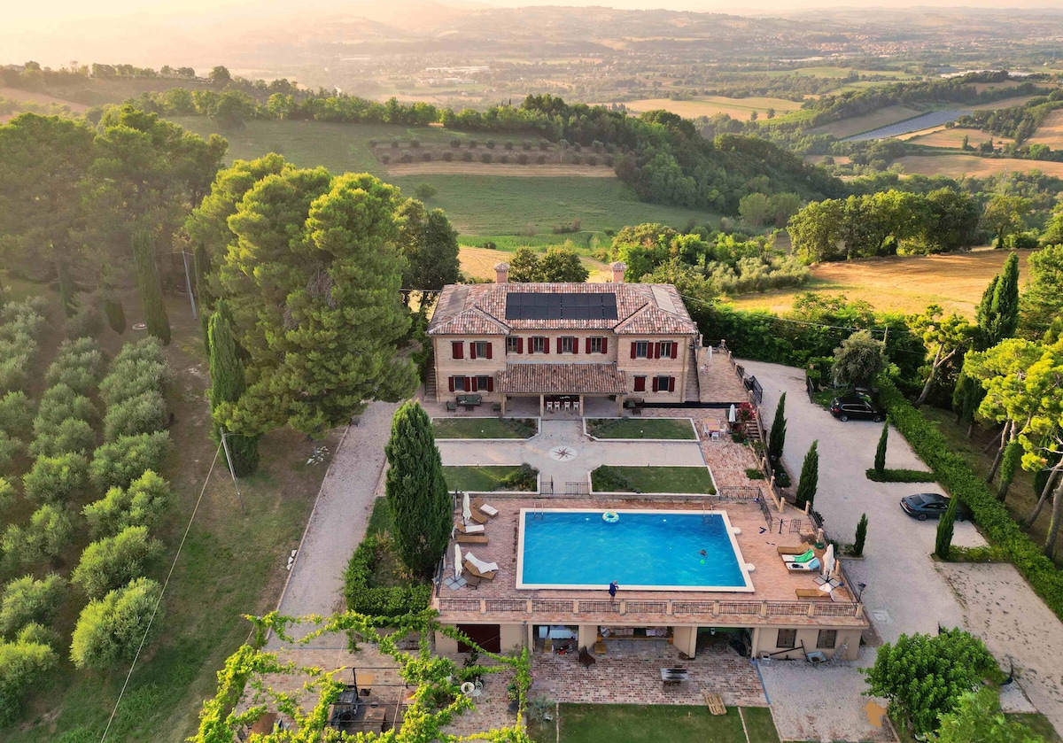 An aerial view of the villa highlights a spacious outdoor area with a swimming pool surrounded by sun loungers. Lush greenery and rolling hills provide a scenic backdrop, while the two-story villa boasts a classic architecture with large windows and a terracotta-colored facade.
