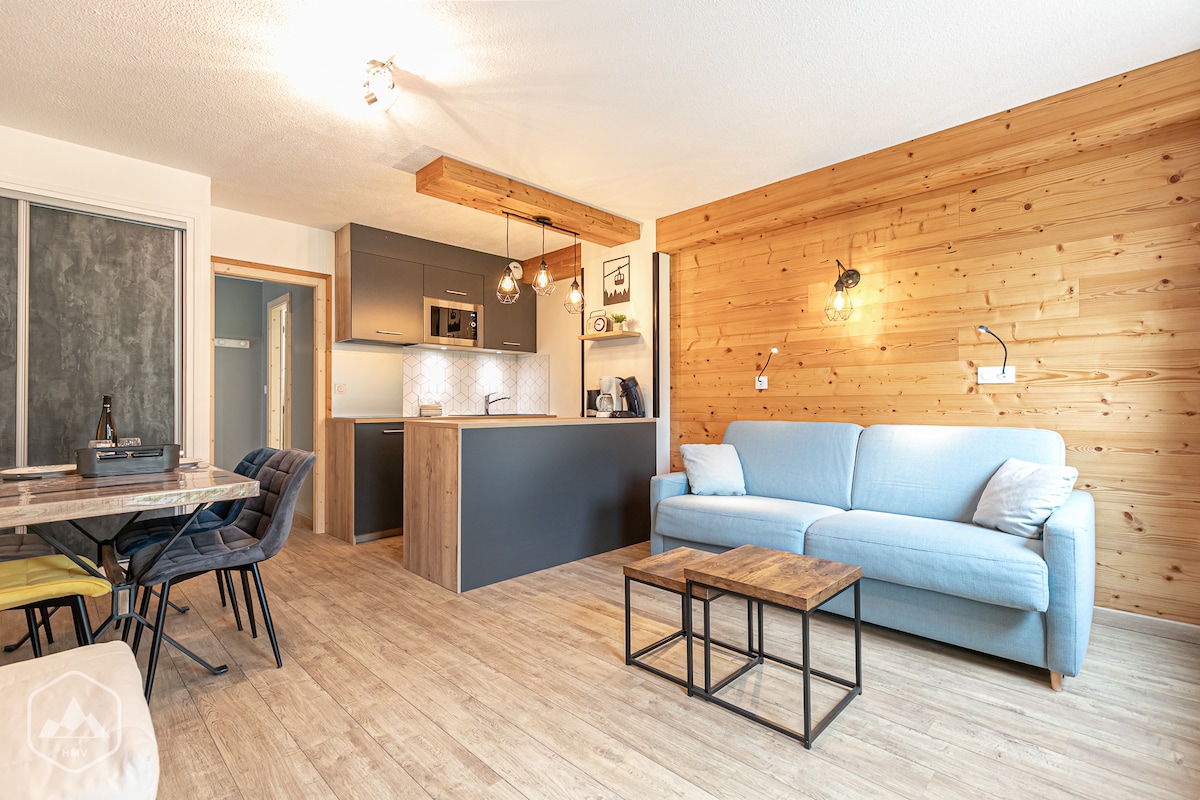 A cozy studio features light wooden walls and modern furnishings. A spacious blue sofa is paired with a wooden coffee table. A dining area with four chairs is adjacent to the kitchenette, which includes sleek cabinetry and modern appliances. Bright, warm lighting enhances the inviting space.