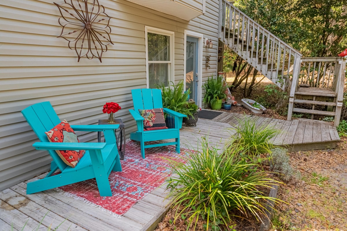 A serene outdoor patio area is featured, showcasing two bright blue chairs positioned on a wooden deck. Decorative cushions adorn the chairs, and a small table is placed between them, complemented by potted plants and foliage enhancing the inviting atmosphere.