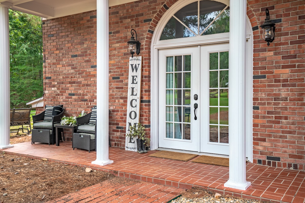 The entrance features double glass doors framed by white pillars, leading to a welcoming porch. A 'Welcome' sign is displayed beside two chairs on a brick floor, complemented by greenery in decorative pots. Natural light illuminates the entryway, inviting guests inside.