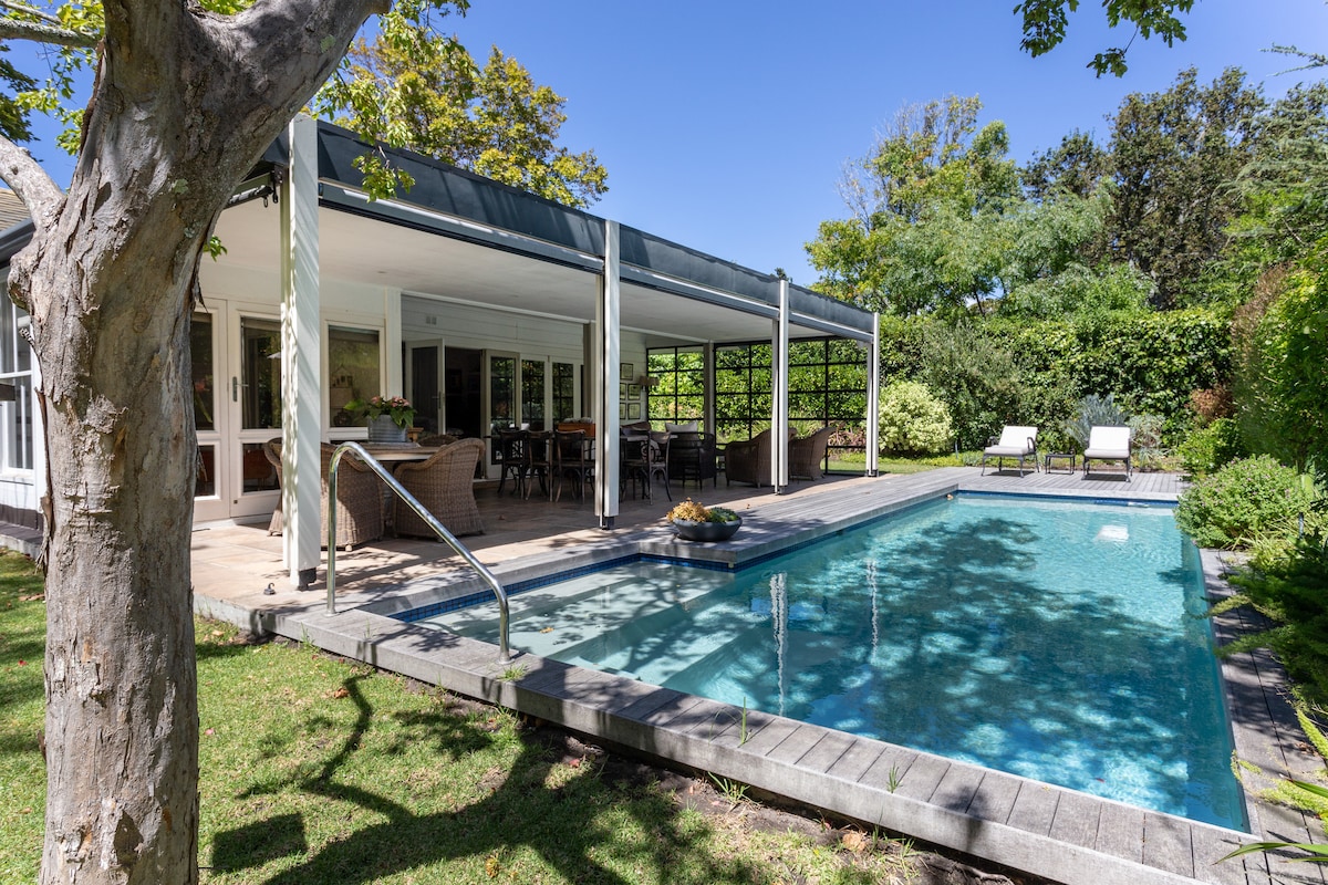 A spacious outdoor area features a swimming pool surrounded by verdant greenery. Comfortable loungers are placed nearby, with a covered patio offering dining and relaxed seating options. Large sliding doors connect the patio to the interior of the home, enhancing the flow between indoor and outdoor spaces.