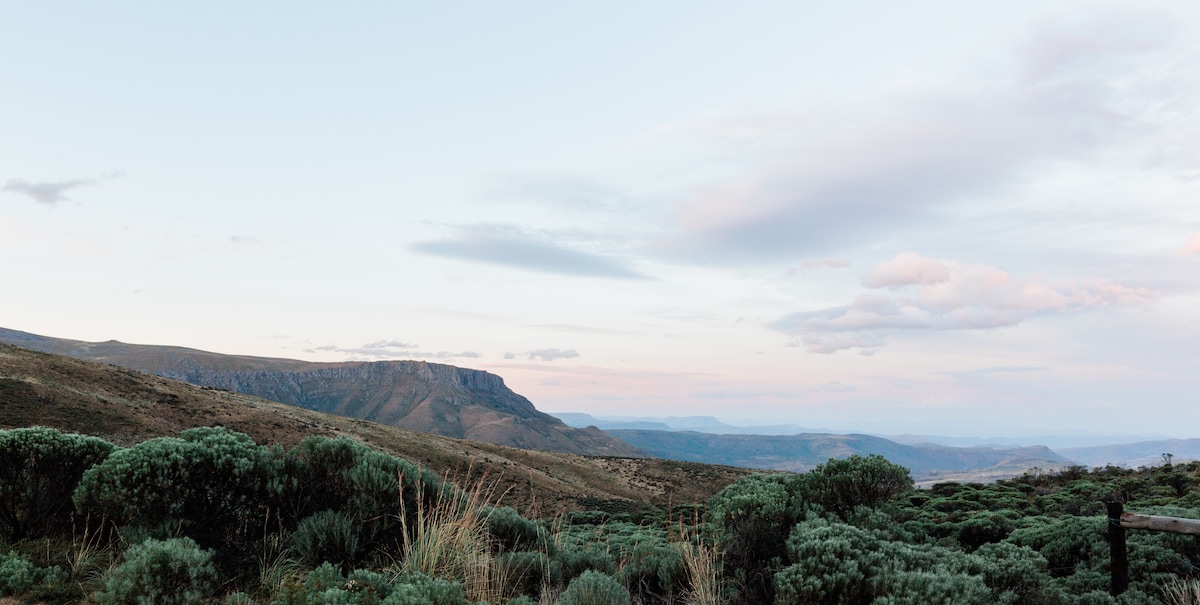 A breathtaking view of the surrounding mountains and valleys is presented, characterized by gentle slopes and a variety of green shrubs. The sky features soft cloud formations, transitioning from blue to pale pastel hues, creating a serene and expansive landscape.