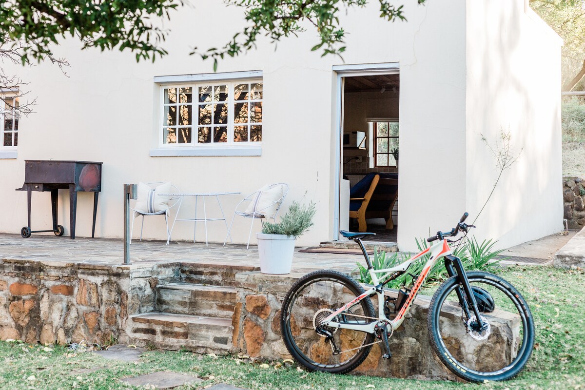 A charming outdoor area features steps leading up to a cottage with a welcoming patio setup. A mountain bike is parked beside a rustic stone wall, and a small grill stands nearby. Natural greenery surrounds the space, creating an inviting atmosphere.