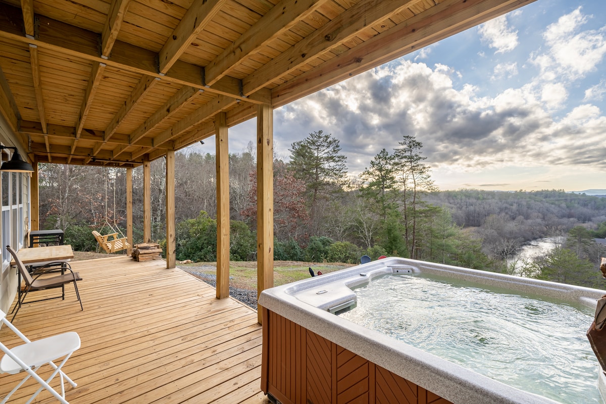 A spacious deck features a hot tub overlooking the Toccoa River, offering scenic views of the surrounding trees and mountains. Wooden flooring complements the natural environment, while nearby lounge chairs provide a comfortable space for relaxation.