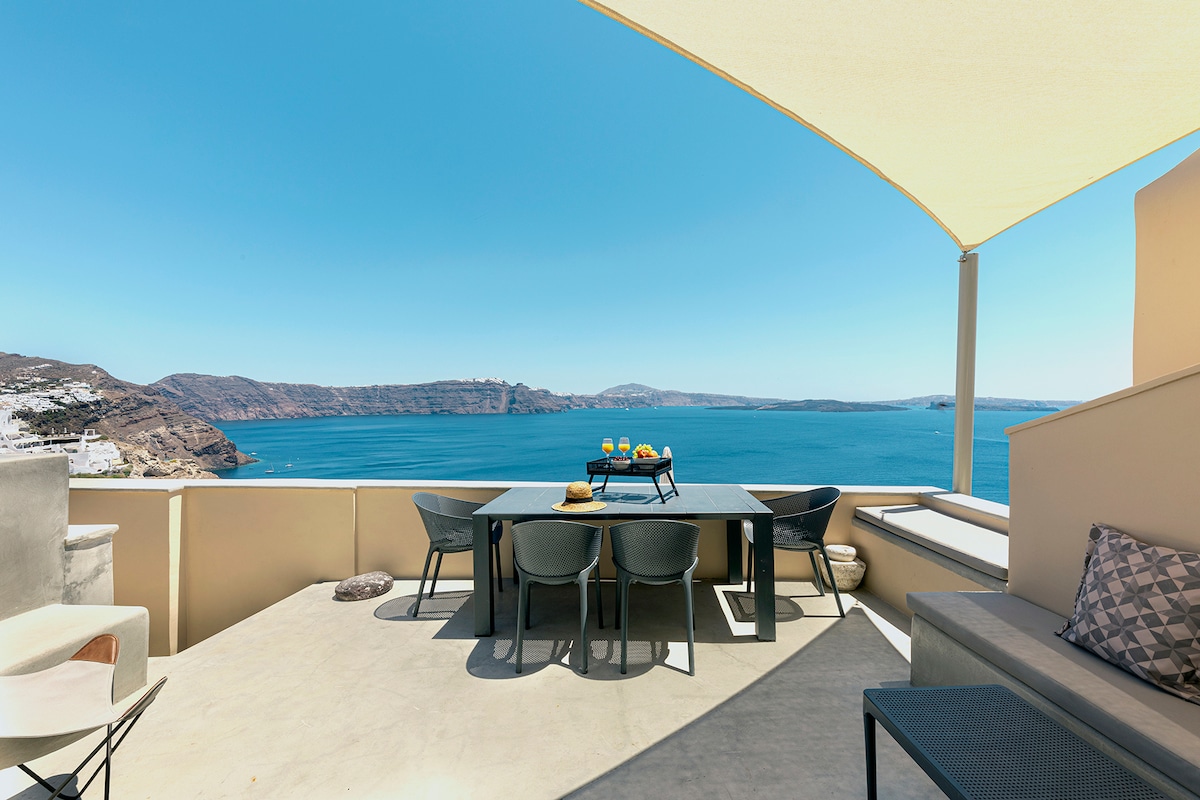 A spacious terrace offers sweeping views of the caldera and the sea, bathed in natural light. Stylish outdoor furniture includes a dining table set for four and a comfortable seating area, all framed by a large sunshade that provides ample shade.