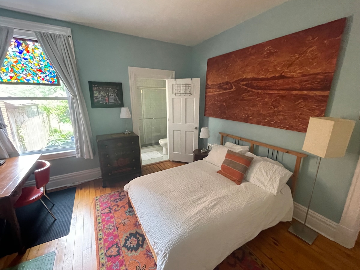 A cozy bedroom features a spacious bed with white linens, situated beside a wooden dresser and a desk. A stained glass window brightens the room, while a private bathroom is visible through a partially open door. A vibrant area rug adds warmth to the wooden floor.