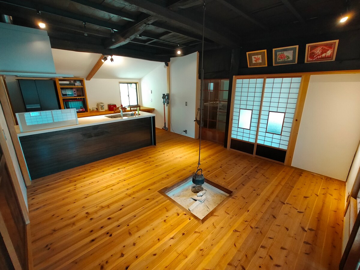 The interior features warm wooden flooring and exposed beams, creating a spacious and welcoming atmosphere. A traditional irori (hearth) is situated at the center, surrounded by sliding shoji doors that separate different areas. Natural light floods the space through the open windows.