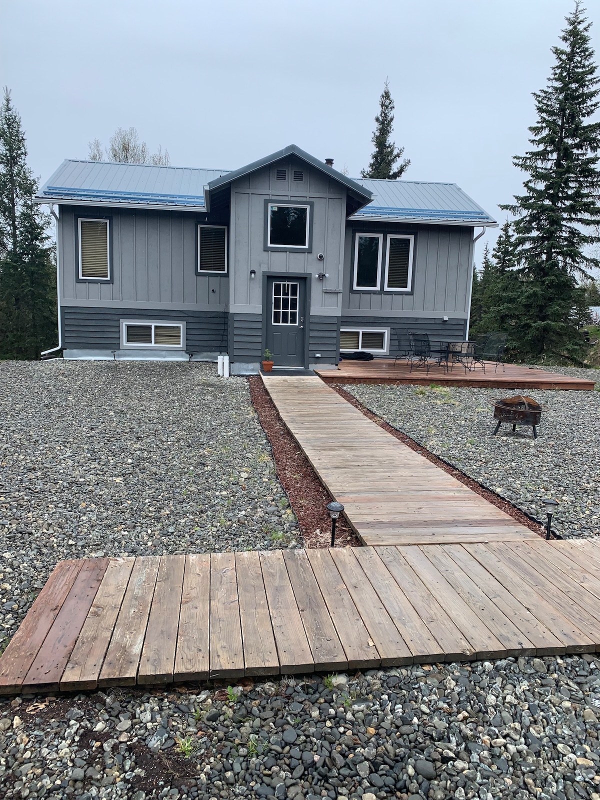 The house features a contemporary exterior with a grey facade and a blue metal roof. A wooden pathway leads to the front door, surrounded by gravel and landscaping. A small fire pit is visible in the yard, with seating arranged nearby for outdoor relaxation.