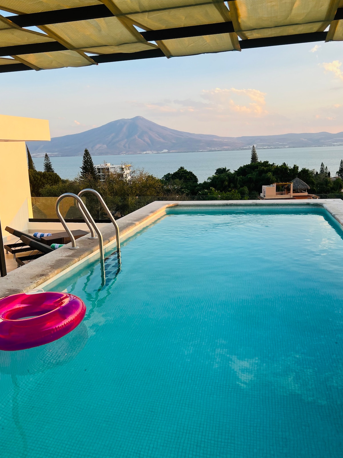 The image captures a bright, inviting swimming pool with clear blue water and a pink inner tube floating on the surface. In the background, a mountain looms over the scenic view of Lake Chapala, while the sky showcases soft pastel colors of a serene sunset.
