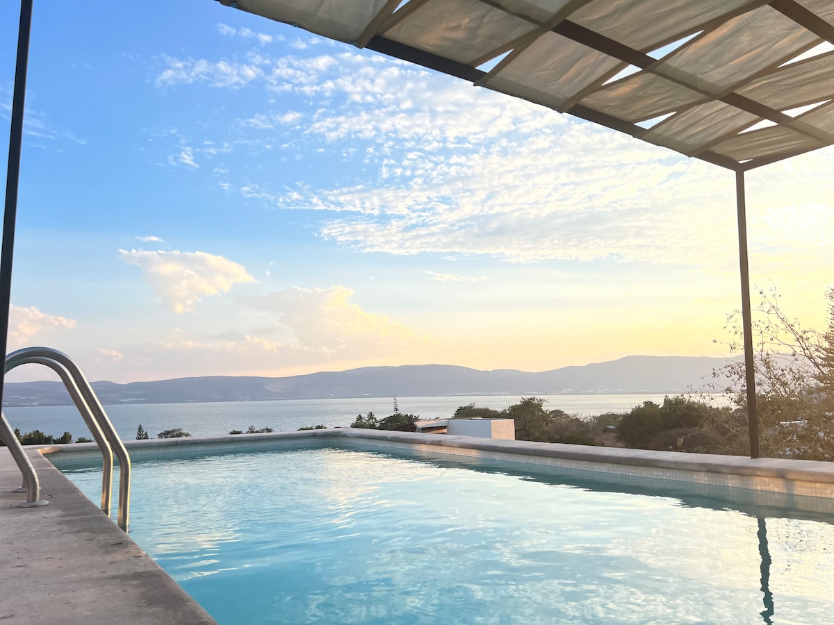 A tranquil swimming pool reflects the soft hues of a sunset, bordered by a covered terrace. The serene landscape of the lake and distant mountains is visible in the background, creating a peaceful atmosphere for relaxation.