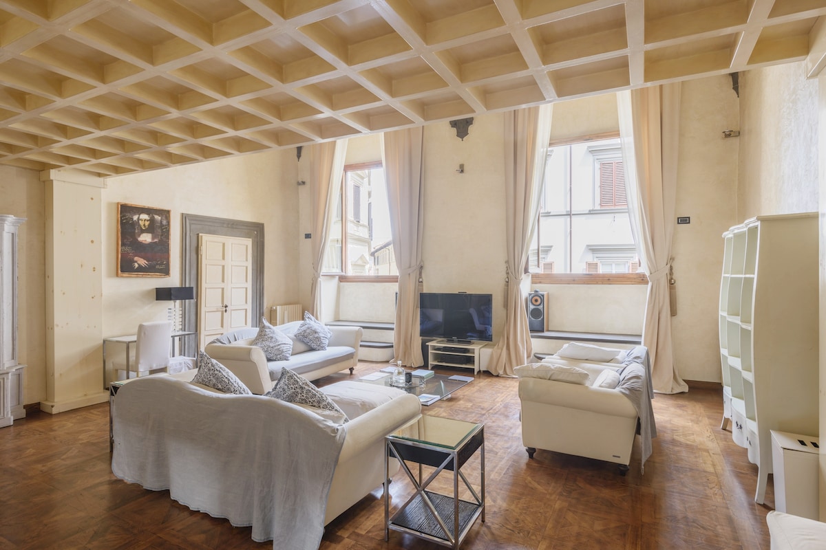 A spacious living area is highlighted by tall windows that allow natural light to fill the room. Comfortable white sofas are arranged around a glass coffee table, and a television is positioned against a wall. Elegant artwork adds a refined touch to the decor.