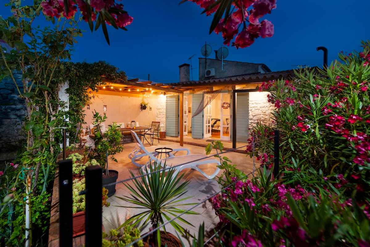 A serene outdoor garden area is framed by vibrant flowers and plants, with comfortable seating arranged under string lights. An inviting patio features a lounge chair and a dining space, creating a harmonious blend of indoor and outdoor relaxation in a peaceful setting.